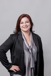 The image shows a woman posing for a portrait. She is wearing a black blazer over a grey top, has dark hair, and is smiling at the camera.