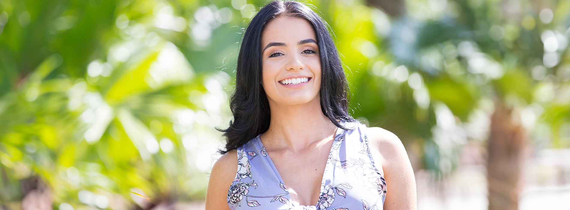 A woman with short hair, smiling and looking directly at the camera, stands outdoors in front of a greenery backdrop.