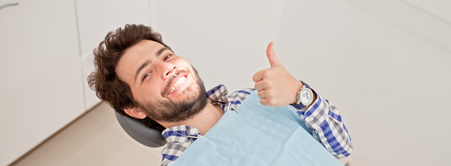 The image shows a man sitting in a dental chair, smiling and giving a thumbs-up gesture.