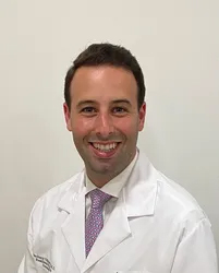 The image shows a man in a white lab coat, smiling at the camera.