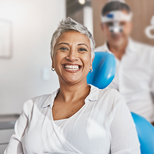 The image features a woman with short hair smiling brightly while seated in an orthodontist s chair, wearing a blue surgical mask and holding a balloon. Behind her is another person, presumably a dental professional, also smiling and looking at the camera. Both are in a modern dental office setting.