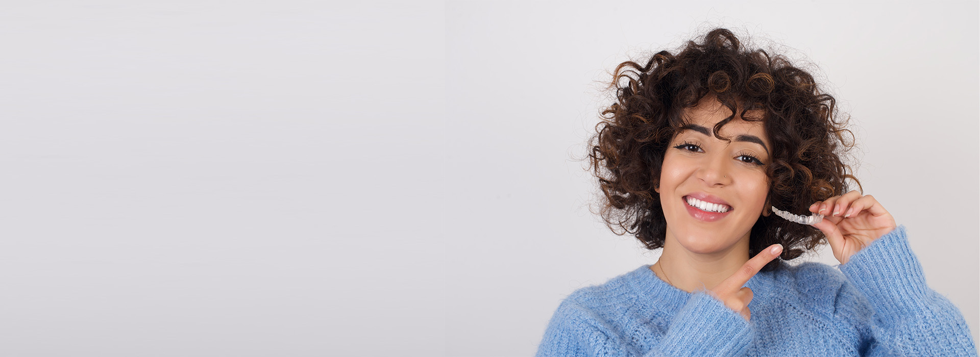 A woman with curly hair is smiling and holding a phone to her ear. She appears to be in a room with a plain wall behind her.