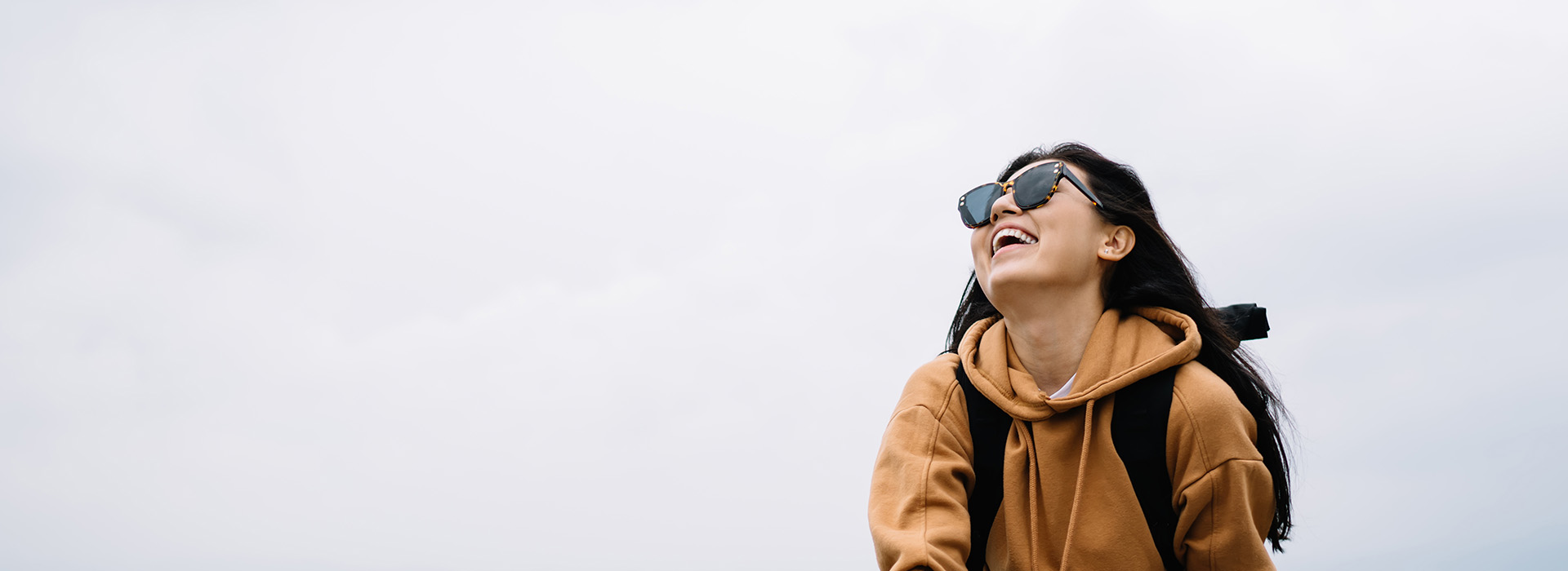 A person is standing outdoors, wearing a backpack and sunglasses, with a smile on their face.