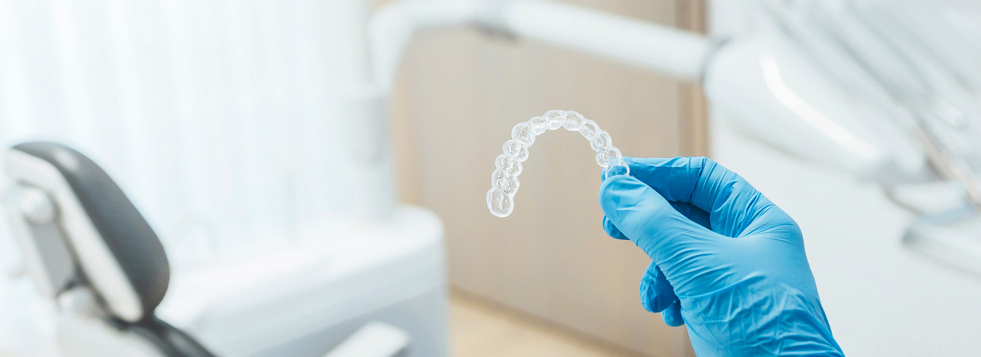 A close-up of a transparent dental impression being held by a gloved hand, with the background showing a dental chair and equipment.