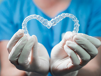 The image shows a pair of clear dental retainers being held by someone wearing white gloves, with the person s hands forming a heart shape in front of them.