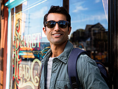 A man in sunglasses and a backpack, smiling at the camera, stands outdoors next to a window with advertising signs.