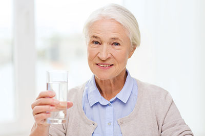 An elderly woman holding a glass of water, smiling and posing for the camera.