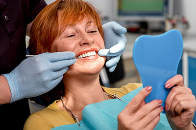 A woman in a dental chair receiving a teeth cleaning, with a hygienist holding a mirror to her mouth.