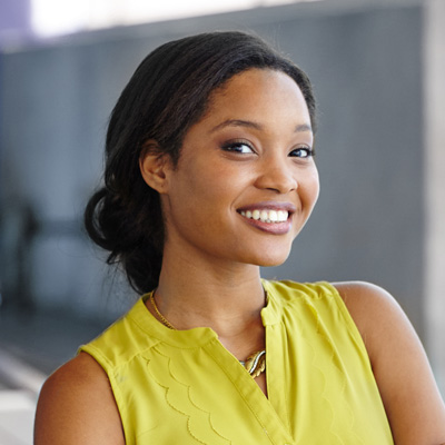 A woman with a radiant smile, wearing a yellow top and standing against a backdrop of a building.
