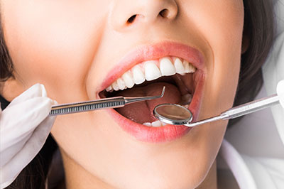 A close-up of a smiling woman with a dental tool in her mouth, wearing gloves and sitting in front of a medical background.