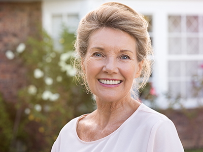 The image shows a woman with short hair, wearing a white top, standing outdoors in front of a house.