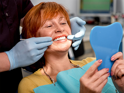 The image shows a woman seated in a dental chair, holding up a blue model of a mouth with teeth. A dentist is standing behind her, wearing gloves and a mask, adjusting the model to fit the woman s mouth.