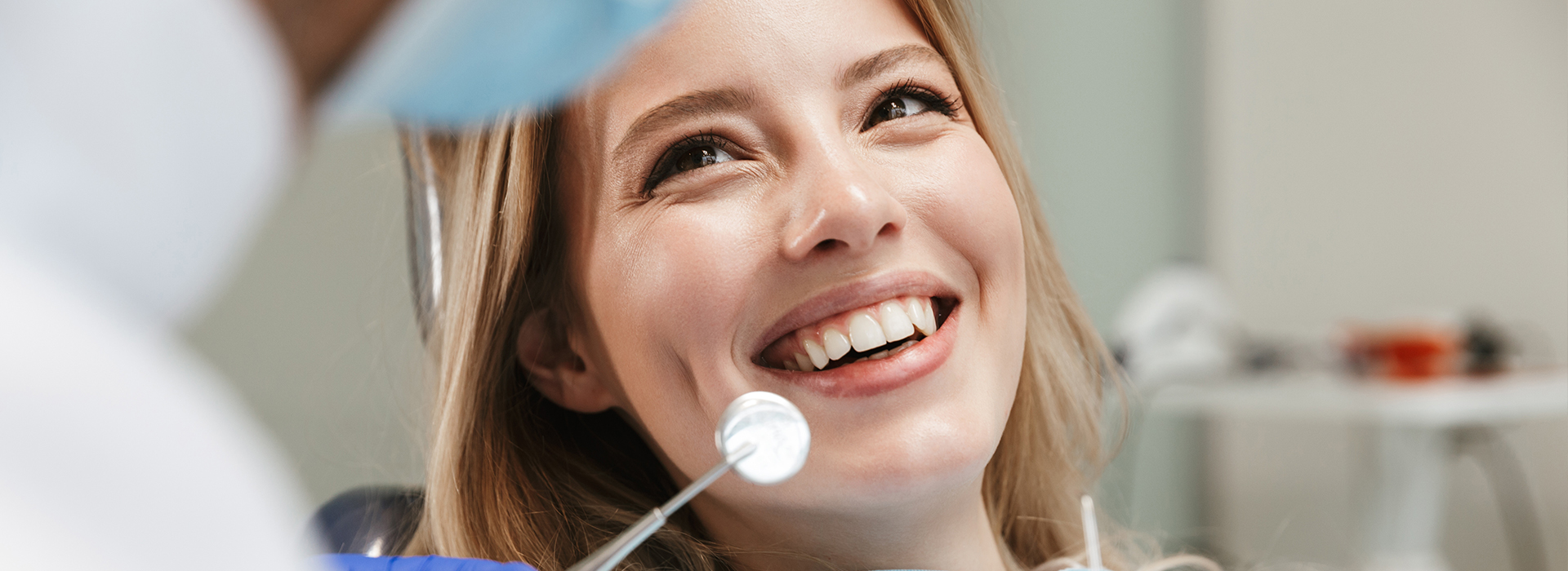 The image shows a woman in a dental chair, smiling at the camera, with a dental professional standing behind her.