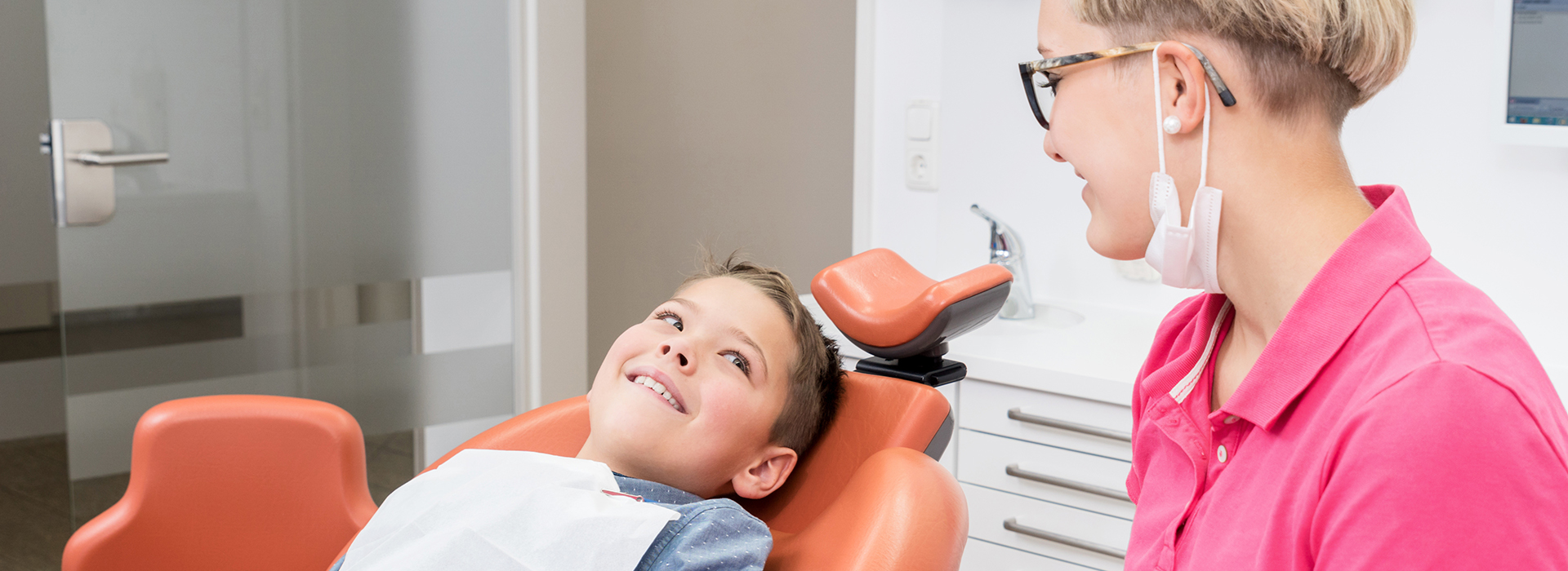 The image depicts a young child sitting in an orange dental chair, receiving dental care from a dentist who is smiling and appears to be assisting the child.