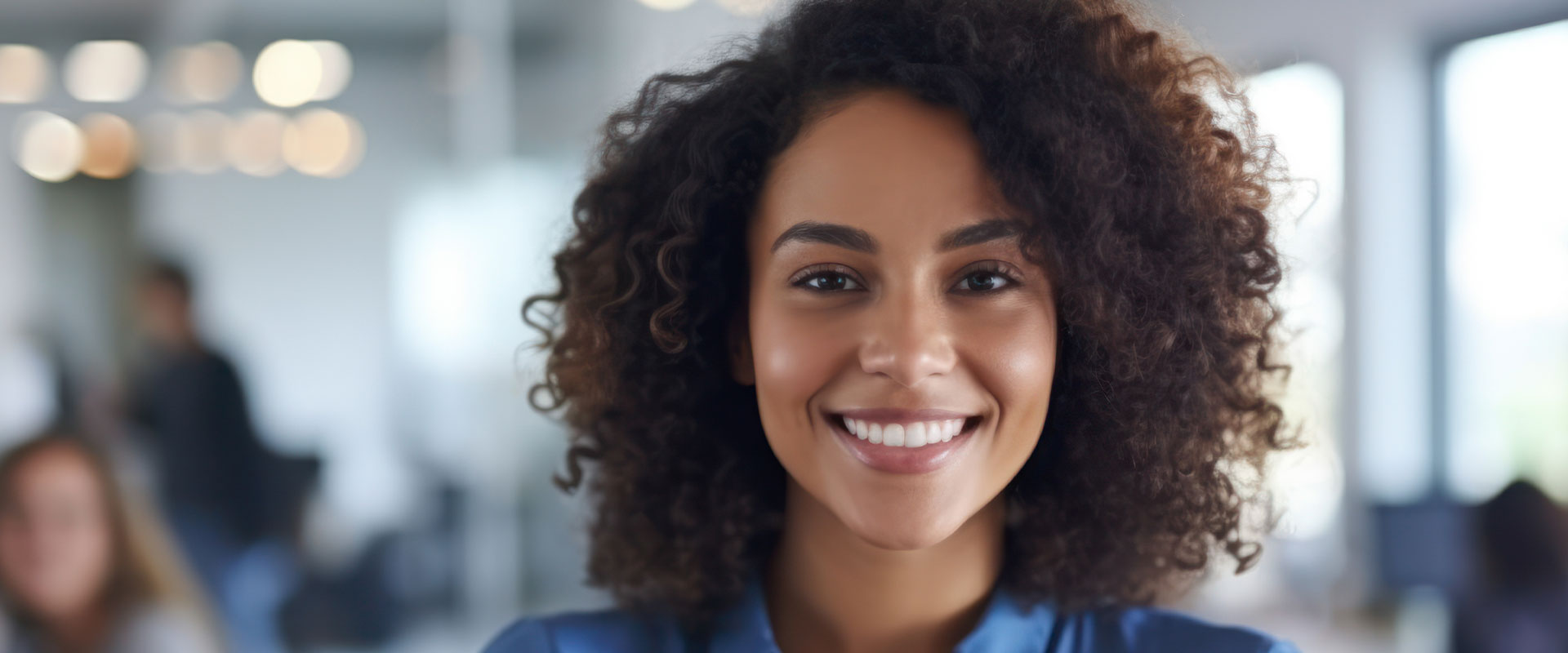 A smiling woman with curly hair in a professional setting, wearing a blue top and standing in front of an office environment.