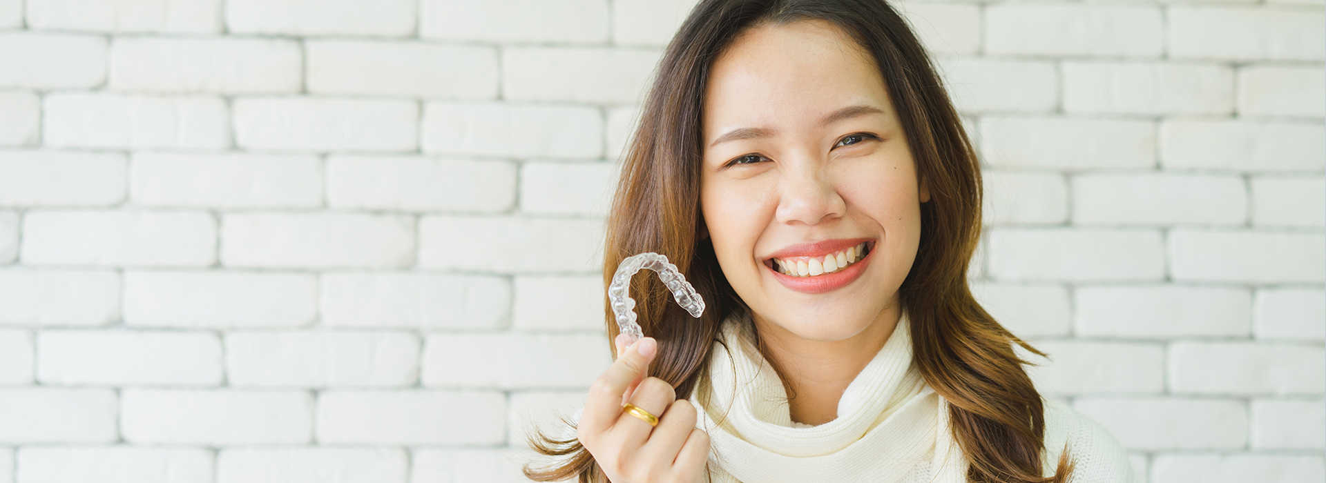 The image shows a woman with a smile, holding up a small object towards the camera. She is wearing a white top and has short hair. The background is blurred but appears to be an indoor setting with a brick wall.