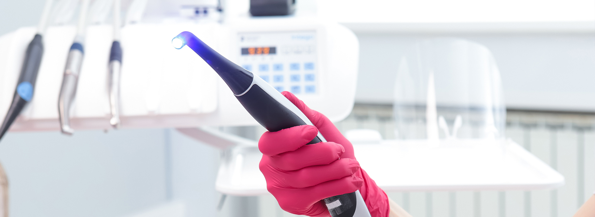 The image shows a person wearing pink gloves and holding a blue device with a black handle, possibly a dental tool or an instrument for some type of procedure, in front of a white background that resembles a medical or dental setting.