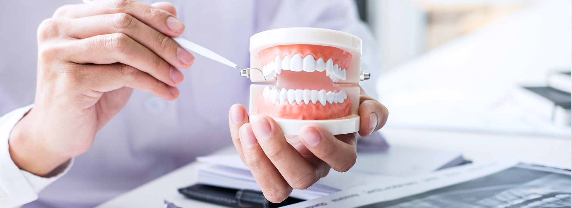 A hand holding a toothbrush in front of a cup filled with dental tools, with a person sitting at a desk in the background.