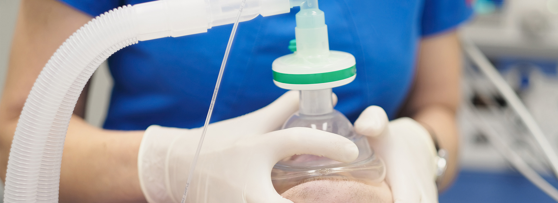 A person in a blue shirt is wearing gloves and holding a transparent bottle with a green lid, possibly containing medication, while standing near medical equipment.