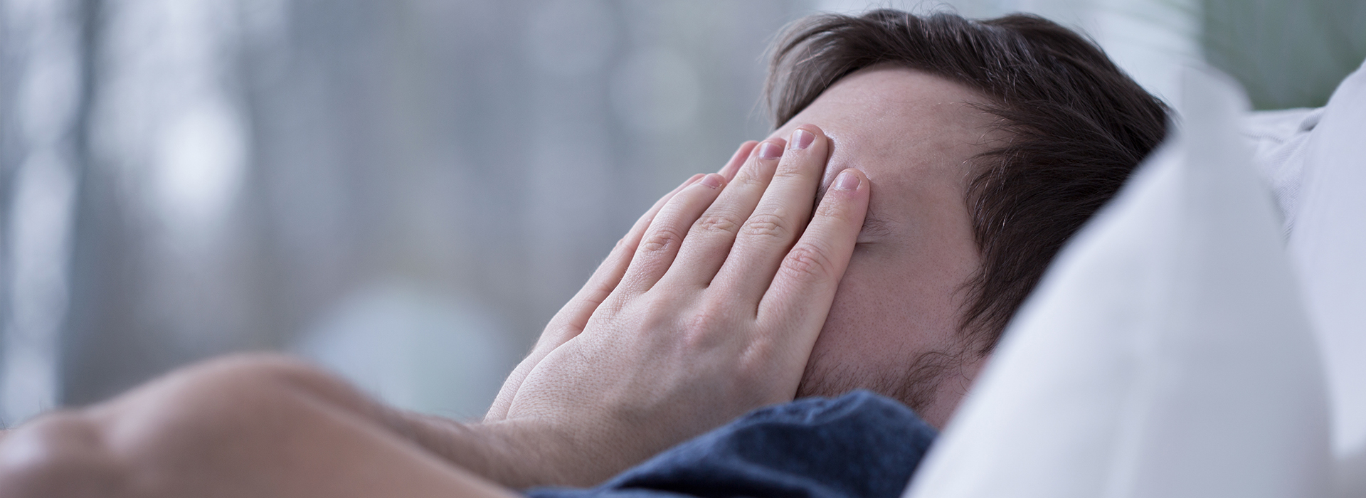 The image shows a man lying in bed with his hands covering his face, which seems to be in a state of distress or disbelief.