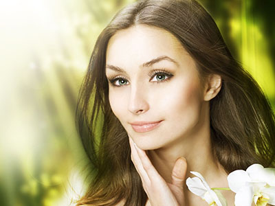 A woman with light skin, long dark hair, and a serene expression is shown against a blurred background of greenery.