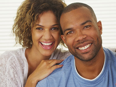 A man and woman smiling at the camera, with the man wearing a short beard and both dressed in casual attire.