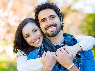 The image shows a man and woman embracing, with the man wearing a blue scarf around his neck and both smiling. They appear to be in an outdoor setting, possibly during autumn or winter, as indicated by the seasonal attire and the foliage in the background.