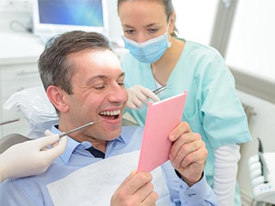 The image shows a man sitting in a dental chair, holding an open envelope with a smile on his face, while a dentist and hygienist stand behind him.