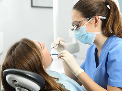 In the image, a dental hygienist is performing a cleaning procedure on a patient s teeth while wearing protective eyewear and a surgical mask.