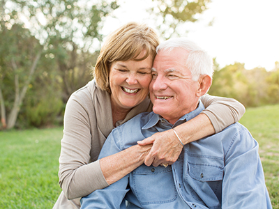 The image shows an elderly couple embracing each other outdoors, with a clear sky and greenery in the background.