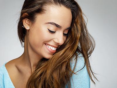 The image shows a woman with long hair, smiling gently and looking downwards. She has fair skin and is wearing a light blue top.