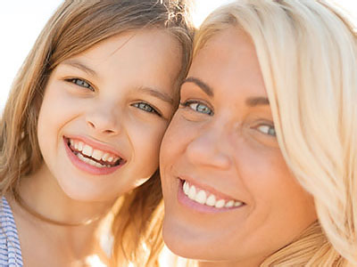 The image shows a woman and a young girl smiling at the camera, with the woman being in focus and the child slightly out of focus.