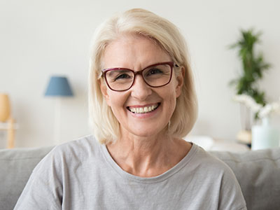 A smiling woman with short blonde hair, wearing glasses and a grey top, is seated in front of a homey background.