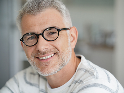 The image is a photograph of a man with short gray hair, wearing glasses and a white shirt. He has a beard and mustache, and he is smiling at the camera.