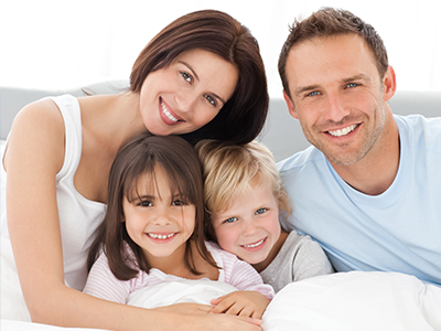 A family of five, including two adults and three children, smiling in a bedroom setting with white bedding.