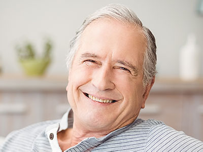 The image shows an elderly man with white hair, wearing a blue shirt, sitting in a chair and smiling.