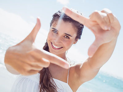 A young woman with long hair is smiling at the camera, holding her fingers up to create a frame around her face. She is wearing a white top and is outdoors on a sunny day.