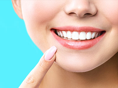 An image of a smiling woman with fair skin, wearing light makeup including pink lipstick and highlighting her cheekbones, set against a solid blue background.