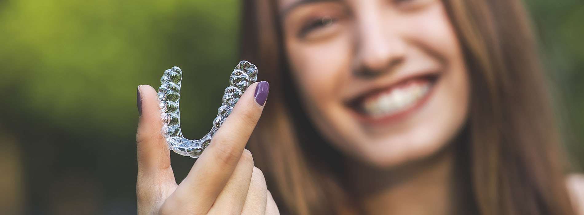 The image shows a smiling person holding a clear dental retainer with their left hand.